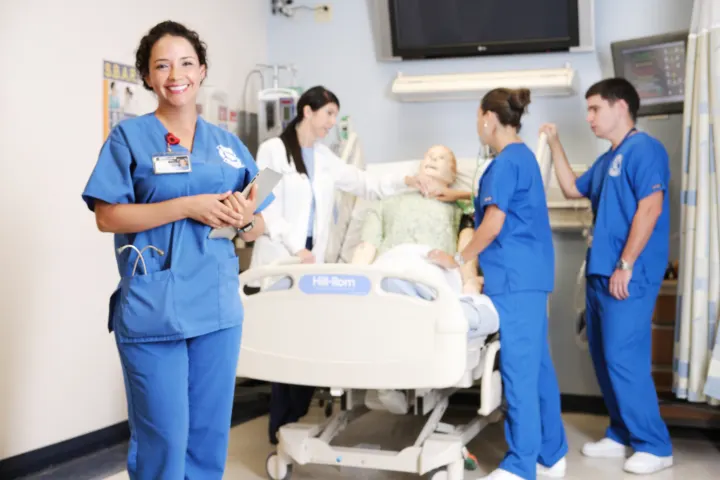 Broward College nurse student posing with clipboard in a patient's room with instructor and nurse student in the background nursing dummy simulator